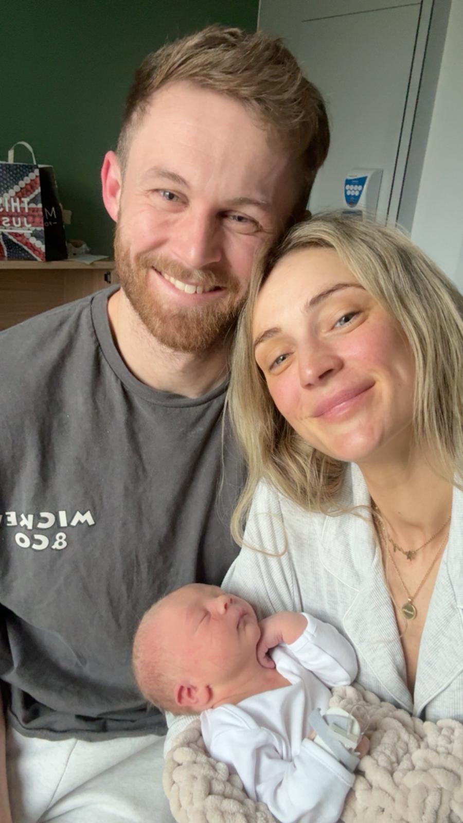 A young couple smiling at the camera, holding a newborn baby