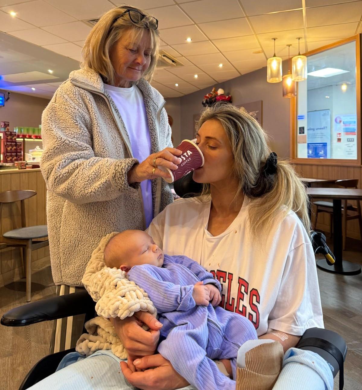 A young woman in a wheelchair holds her infant son while her mother holds a cup for her to drink coffee