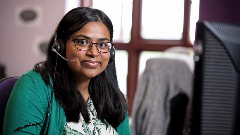 Stroke Association Support Line Officer, Hema, sitting at her desk, with her telephone headset on, smiling. 
