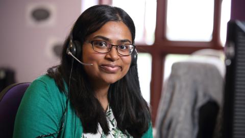Hema, from our Stroke Support Helpline, sits, smiling, at her desk with her telephone headset on as if waiting to answer a call.