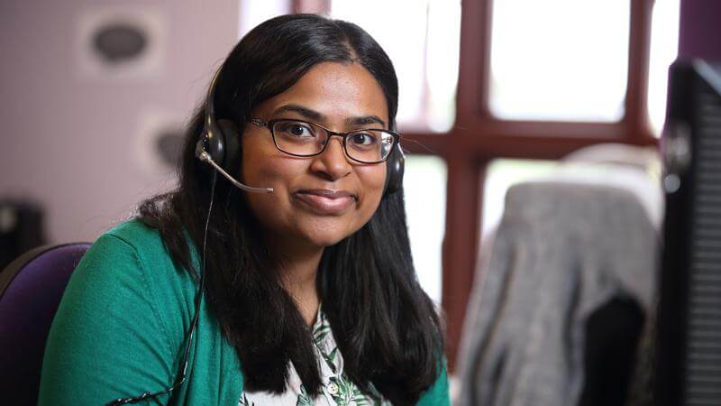Hema, from our Stroke Support Helpline, sits, smiling, at her desk with her telephone headset on as if waiting to answer a call.