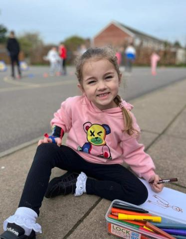 Jess, a childhood stroke survivor, sits on the pavement colouring and smiling