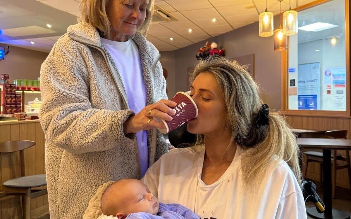 A young woman holds a baby while her own mother helps her drink a coffee