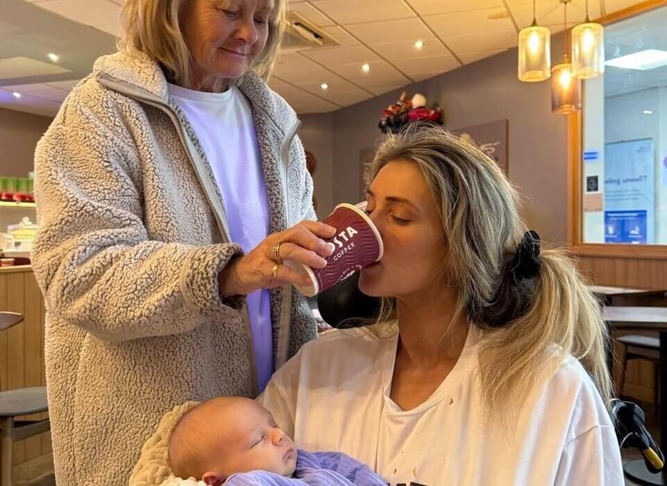 A young woman holds a baby while her own mother helps her drink a coffee