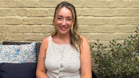 Stroke survivor, Leighton, sitting on a bench in her garden in front of a painted brick wall, looking at the camera and smiling. 