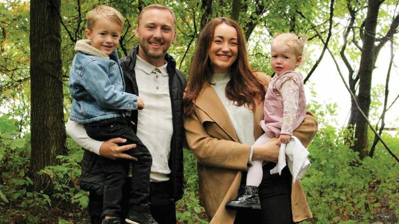Stroke survivor, Louise, stands smiling with her family. She is holding her young daughter, while her husband, who is stood next to her, holds their young son.