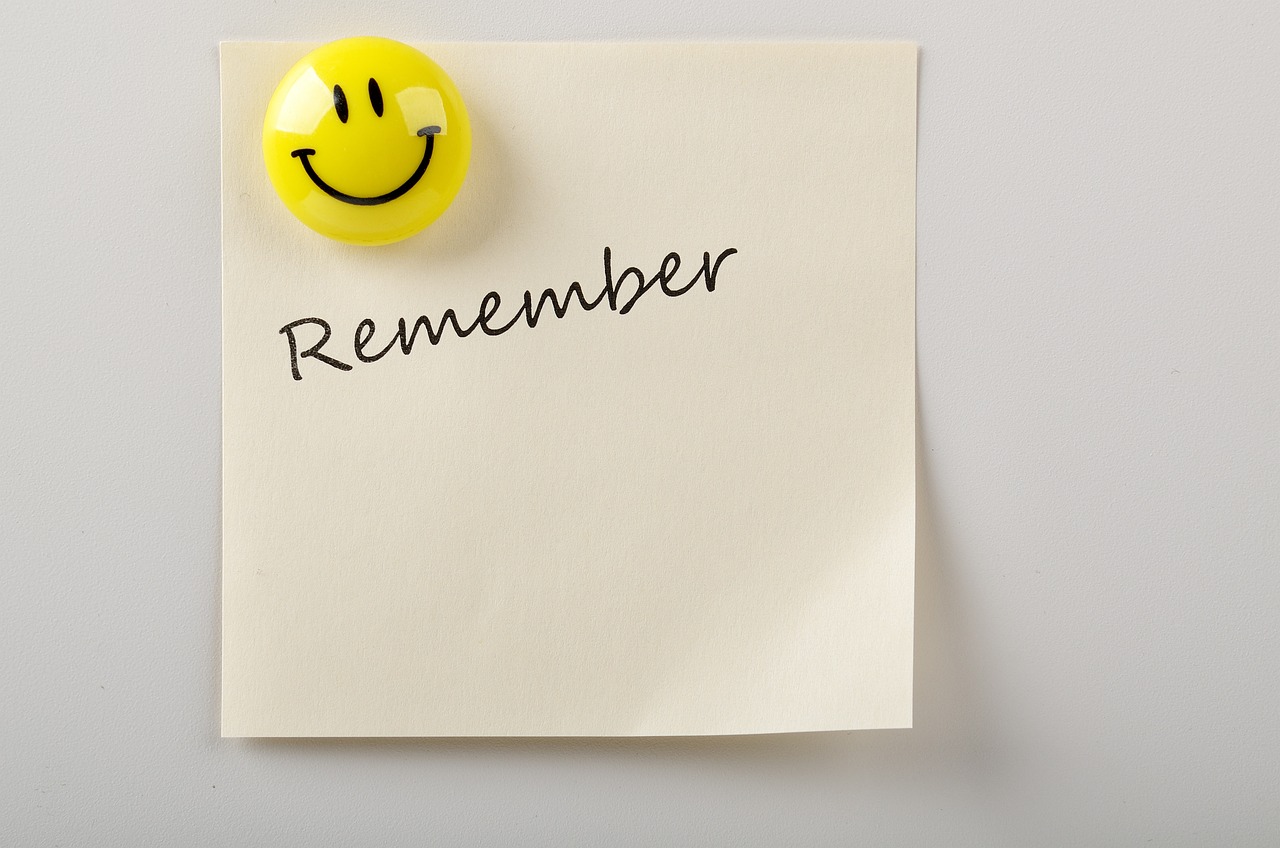 A photo of a yellow smiley face magnet, holding up a piece of paper on a white surface. On the paper, it says 'Remember'.
