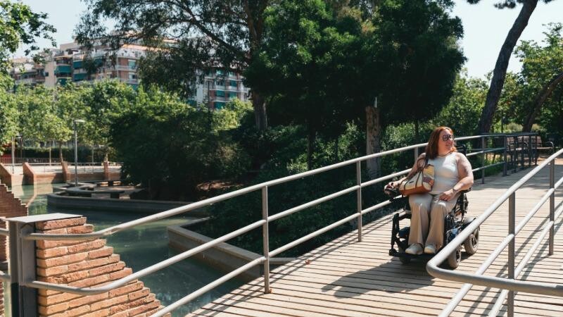 A stock image of a lady in a mobility scooter riding over a bridge on a sunny day. 