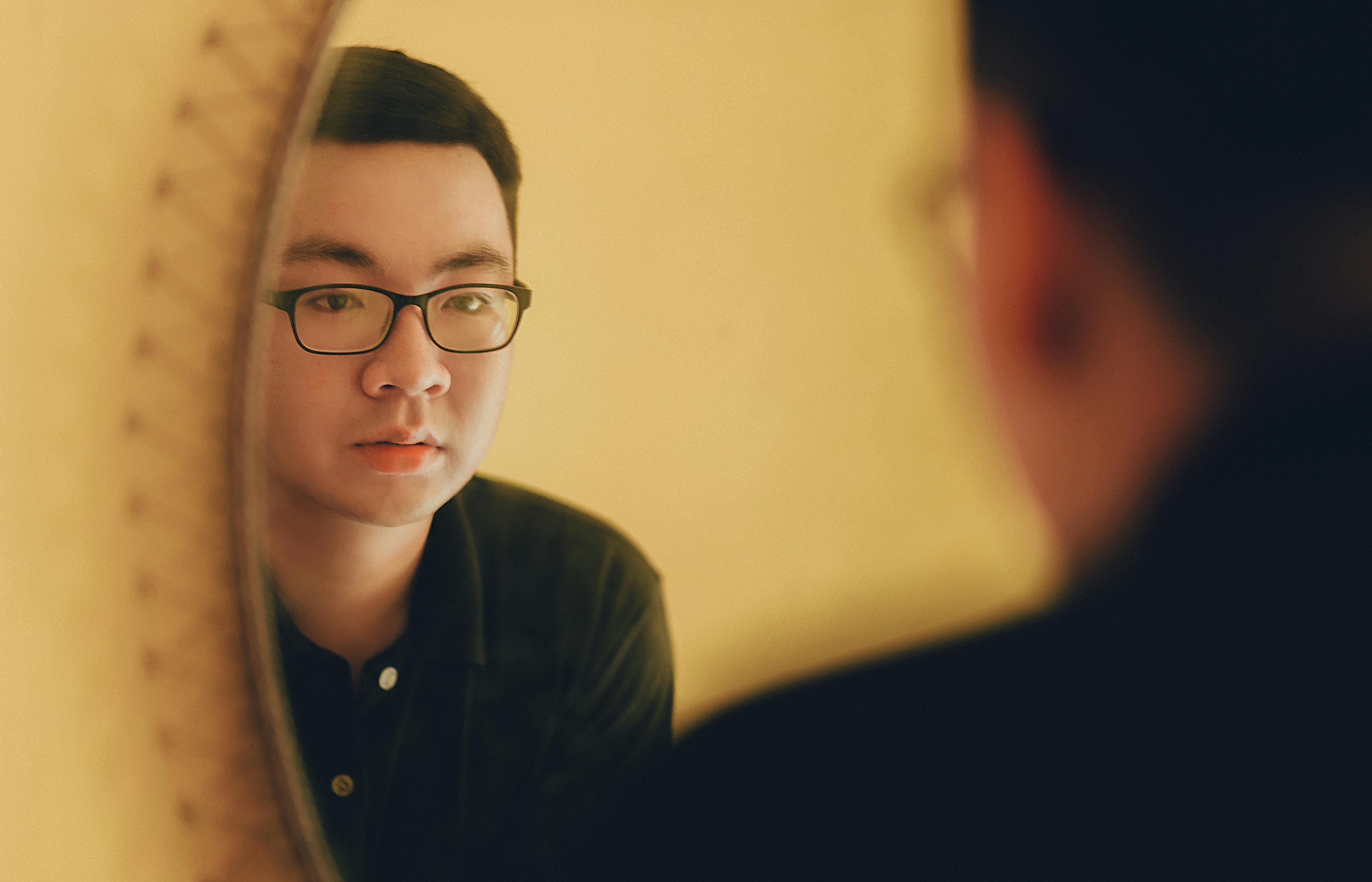 A photo is taken over the should of a man wearing glasses, looking at his reflection in the mirror.