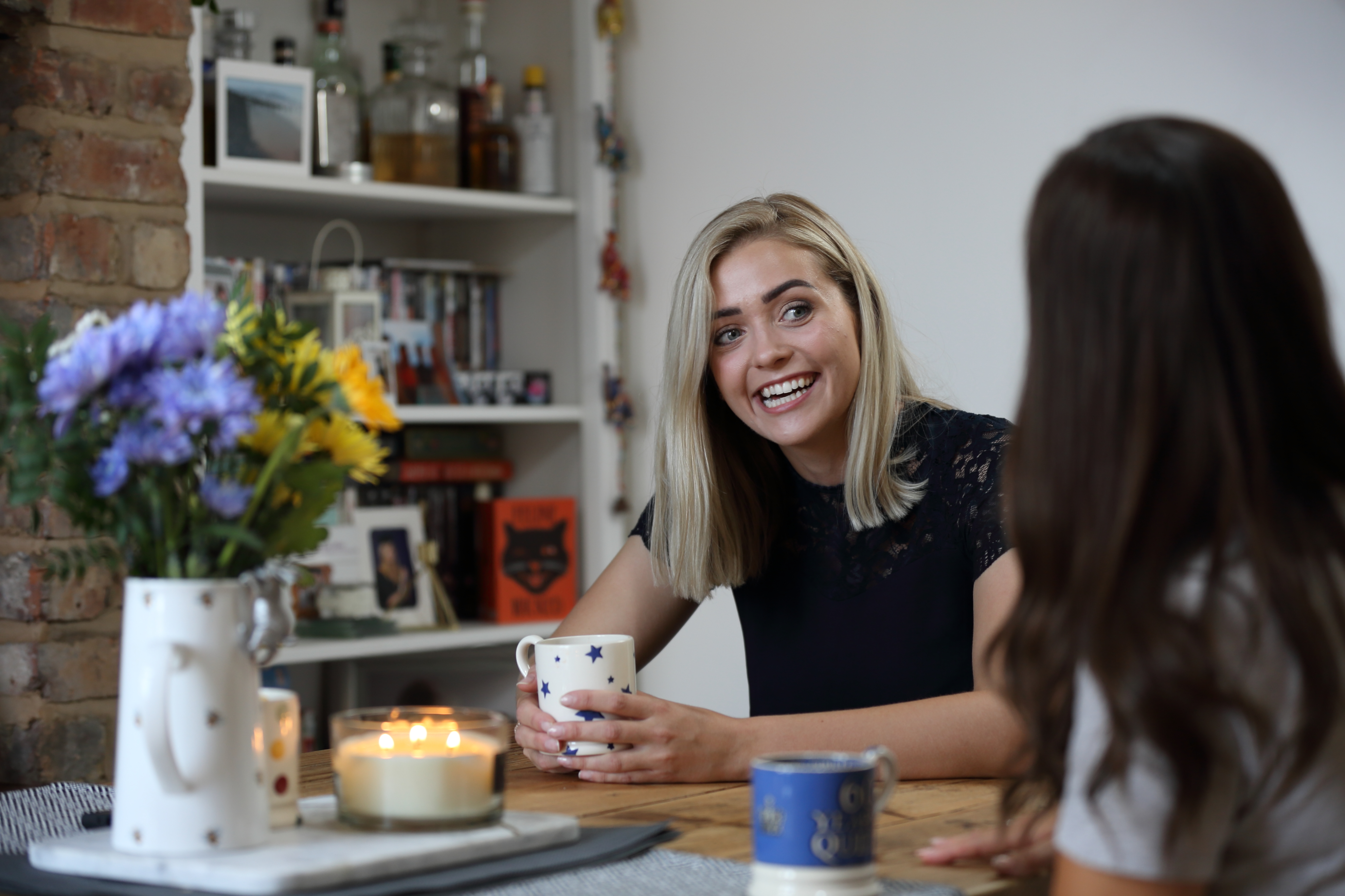 Two women are sat having cup of coffee in a relaxed environment, one is laughing looking at the other one.