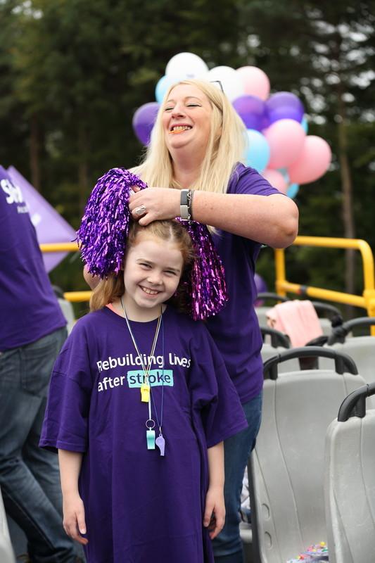 A woman smiles as she puts pompoms in a young girls hair, they are both wearing Stroke t-shirts
