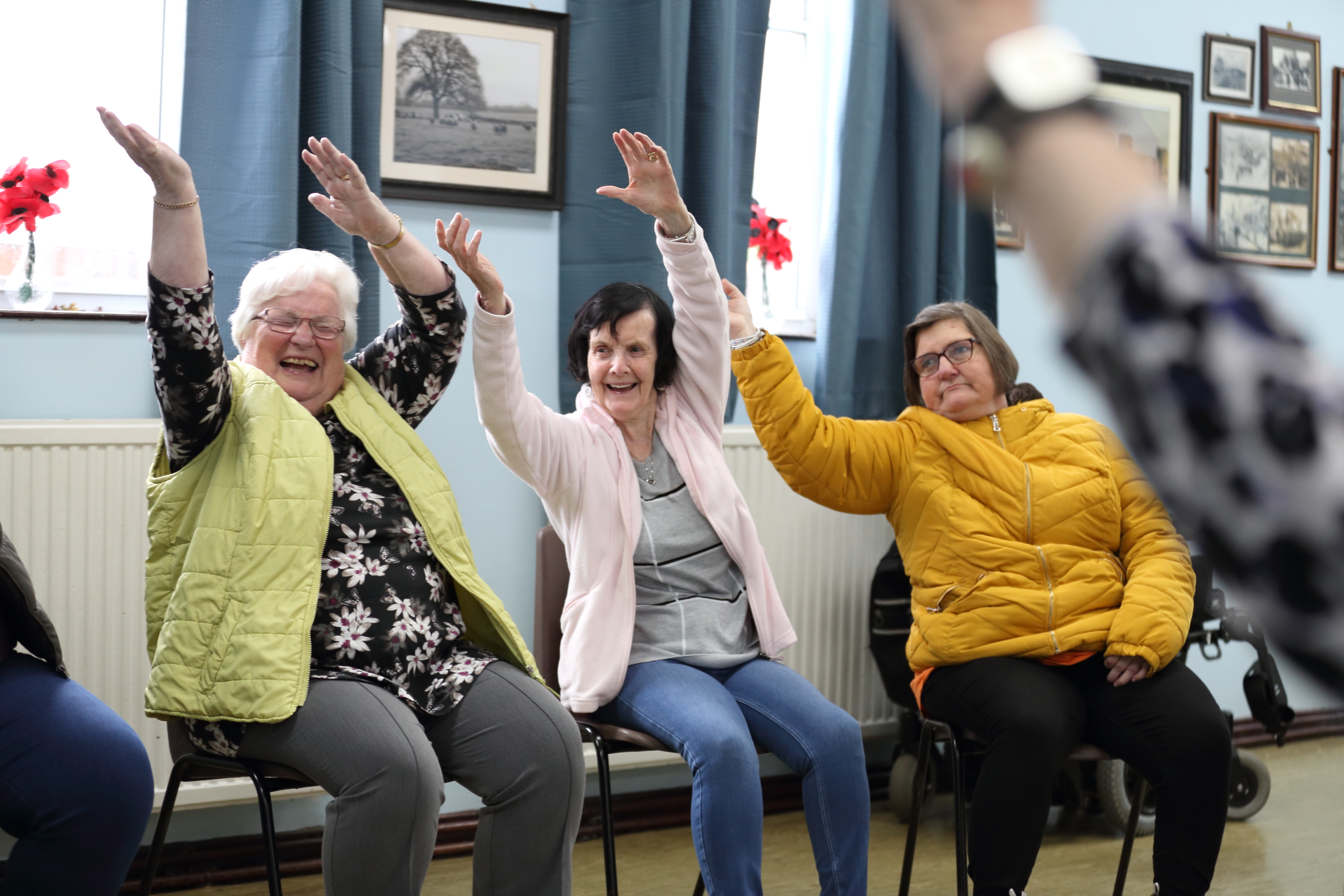 A group of stroke survivors at a group meeting, are all laughing and waving their hands in the air.