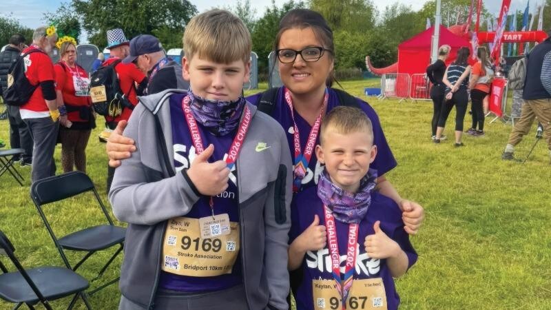 Carer, Shannon, stands with two of her younger brothers, smiling at the finish line of a fundraising run they did together. They're all wearing medals and purple Stroke Association t-shirts.
