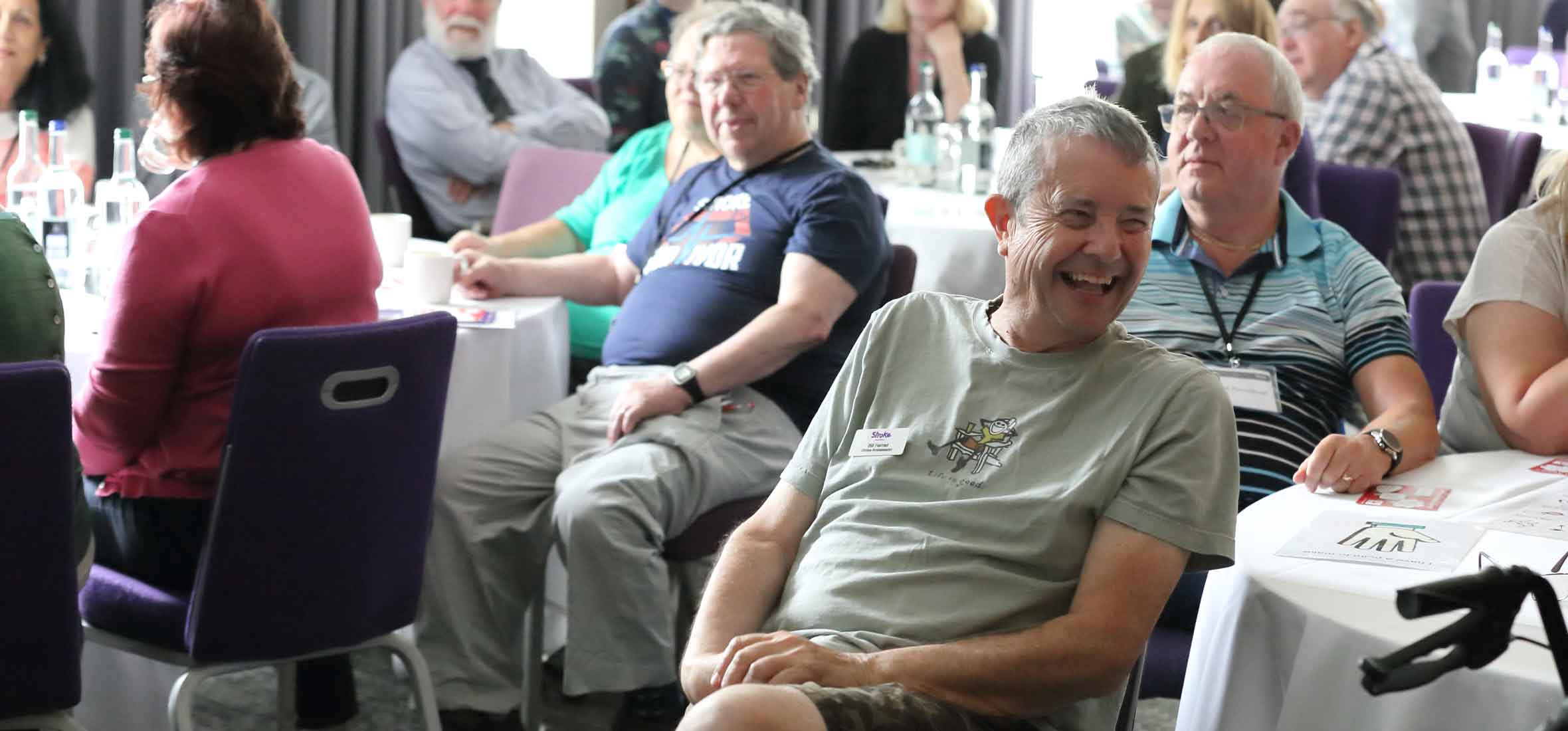 A man looking to the right and laughing, amidst a large room full of people.
